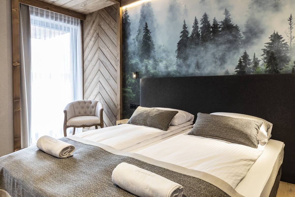Cama doble con dos almohadas, una silla y un amplio mural temático de bosque en una habitación de hotel.