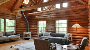 A log cabin interior with a high ceiling, wooden beams, a fireplace, a sofa, a coffee table, and a lamp. - 11,000sqft sleeps 29, w/ POOL! private lake, hiking trails, hot tub, 130 acres! (Logan)