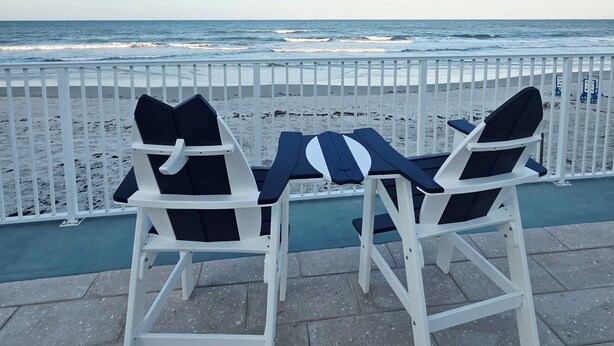 Two white lounge chairs with black cushions, a small table, and a view of the ocean through a white fence. - Ocean View Studio • Steps to Beach (Daytona Beach)