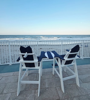 Two white lounge chairs with black cushions, a small table, and a view of the ocean through a white fence.