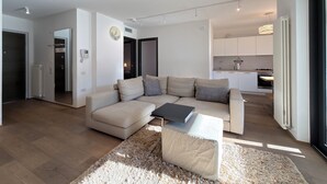 A modern living room with a beige sofa, a coffee table, and a fluffy rug. There is a kitchen area with white cabinets and a dining table. - Apartment with a beautiful terrace (Riva del Garda)