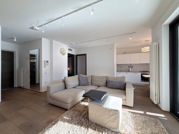 Een moderne woonkamer met een beige bank, een salontafel en een zacht tapijt. Er is een keuken met witte kasten en een eettafel.
