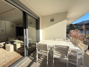 Een moderne woonkamer met een glazen deur die leidt naar een balkon. Het balkon heeft een witte tafel, stoelen en een bloempot.