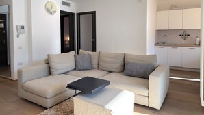 A modern living room with a beige sectional sofa, a small black coffee table, and a fluffy rug. There is a kitchen area with white cabinets and a sink visible through an open doorway. - Apartment with a beautiful terrace (Riva del Garda)