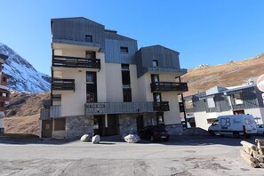 Um edifício de vários andares com varandas, uma placa com a inscrição "Le Plein Soleil" e uma área de estacionamento com uma van e toras.