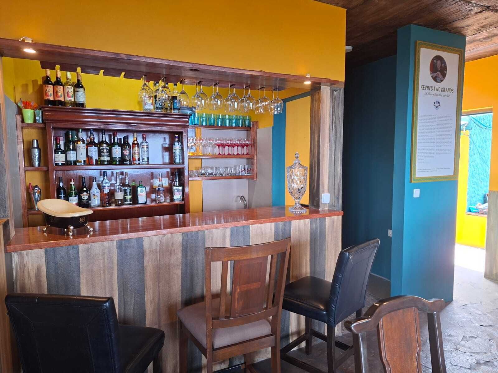 A bar area with a wooden counter, shelves with bottles, glasses, and a framed picture on the wall.