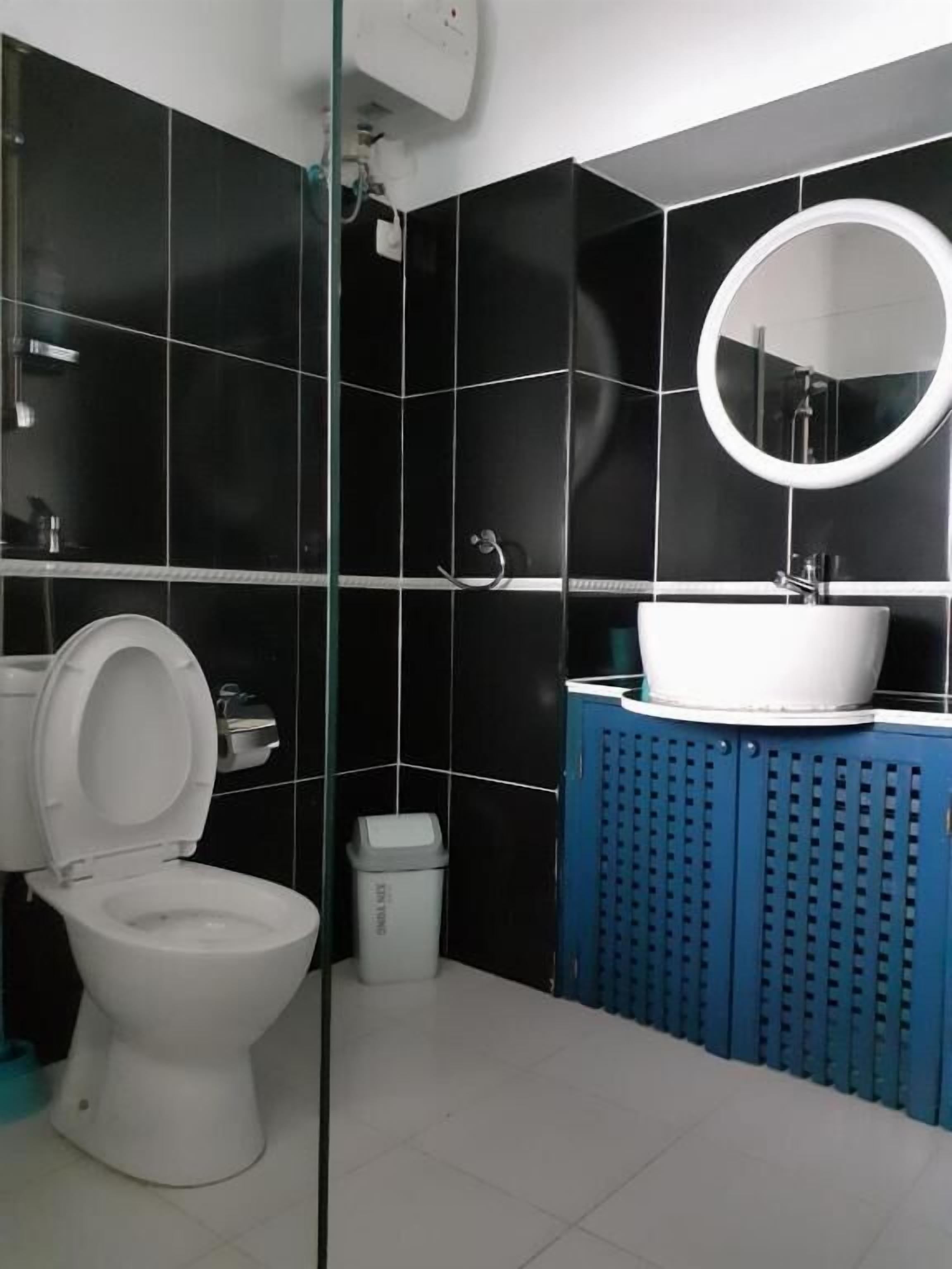 A modern bathroom with black tiles, a white toilet, a blue cabinet, and a round mirror.