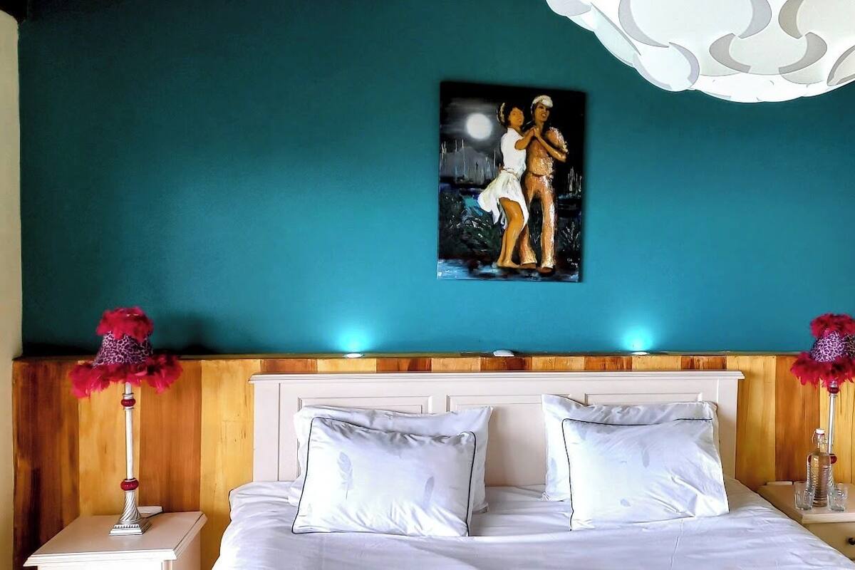 Quarto com parede verde-água, uma pintura de duas figuras, roupa de cama branca e luminárias decorativas.