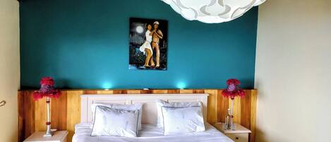 A bedroom with a teal wall, a painting of two figures, white bedding, and decorative lamps.