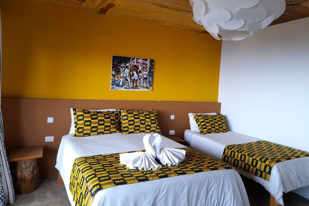 Quarto de hotel com duas camas, cada uma com colcha e almofadas estampadas, cabeceira de madeira, um quadro na parede e uma luminária de teto branca.