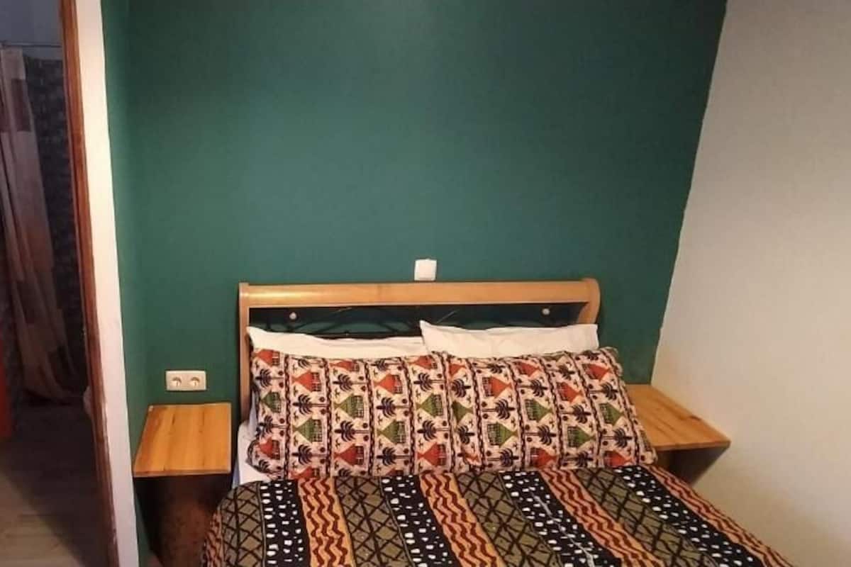 Quarto com cama, cabeceira de madeira, mesa de cabeceira e luminária fixa na parede.