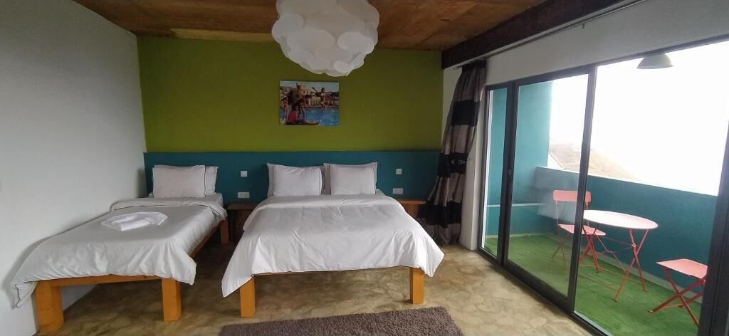 A room with two beds, a green wall, a painting, and a sliding glass door to a balcony with red chairs.