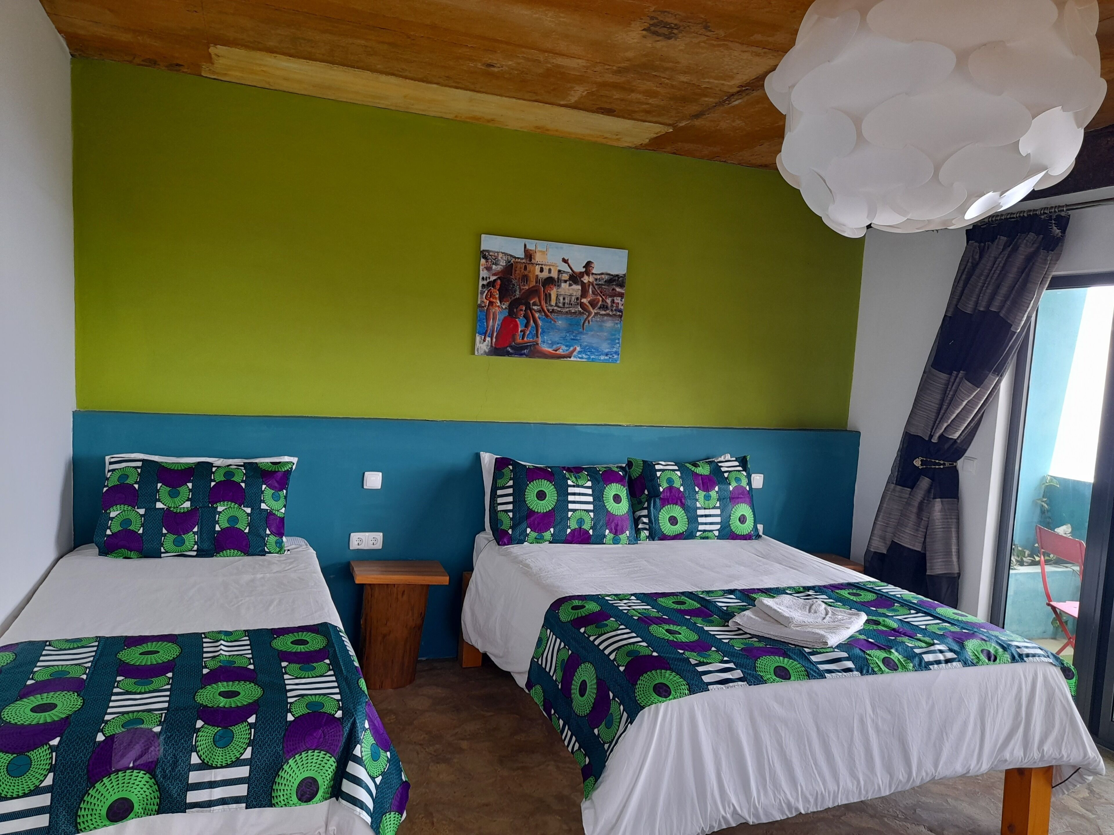 A room with two beds, each with a patterned bedspread and pillows, a small wooden nightstand, a wall-mounted painting, and a large window with curtains.
