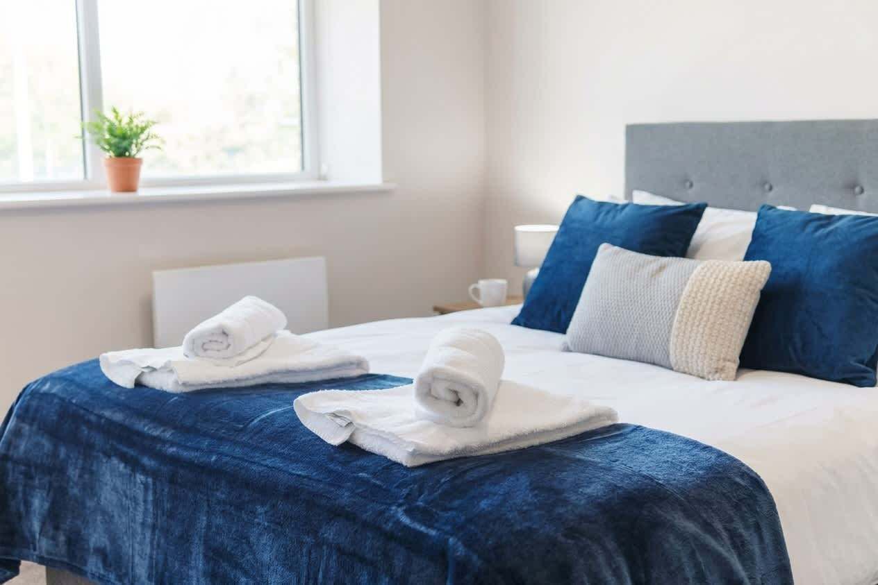 Un lit bien fait, avec une couvre-lit bleue, deux serviettes blanches roulées et une tête de lit grise.