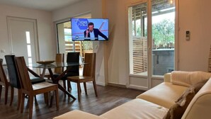 Ein Wohnzimmer mit einem Glastisch, Holzstühlen, einem beigefarbenen Sofa und einem Fernseher, der eine Nachrichtensendung zeigt.