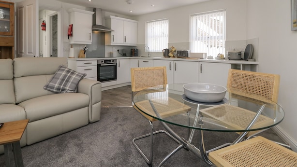 A modern kitchen with white cabinets, a built-in oven, and a dining area with a glass table and wicker chairs. - Betty's Loft (Ambleside)