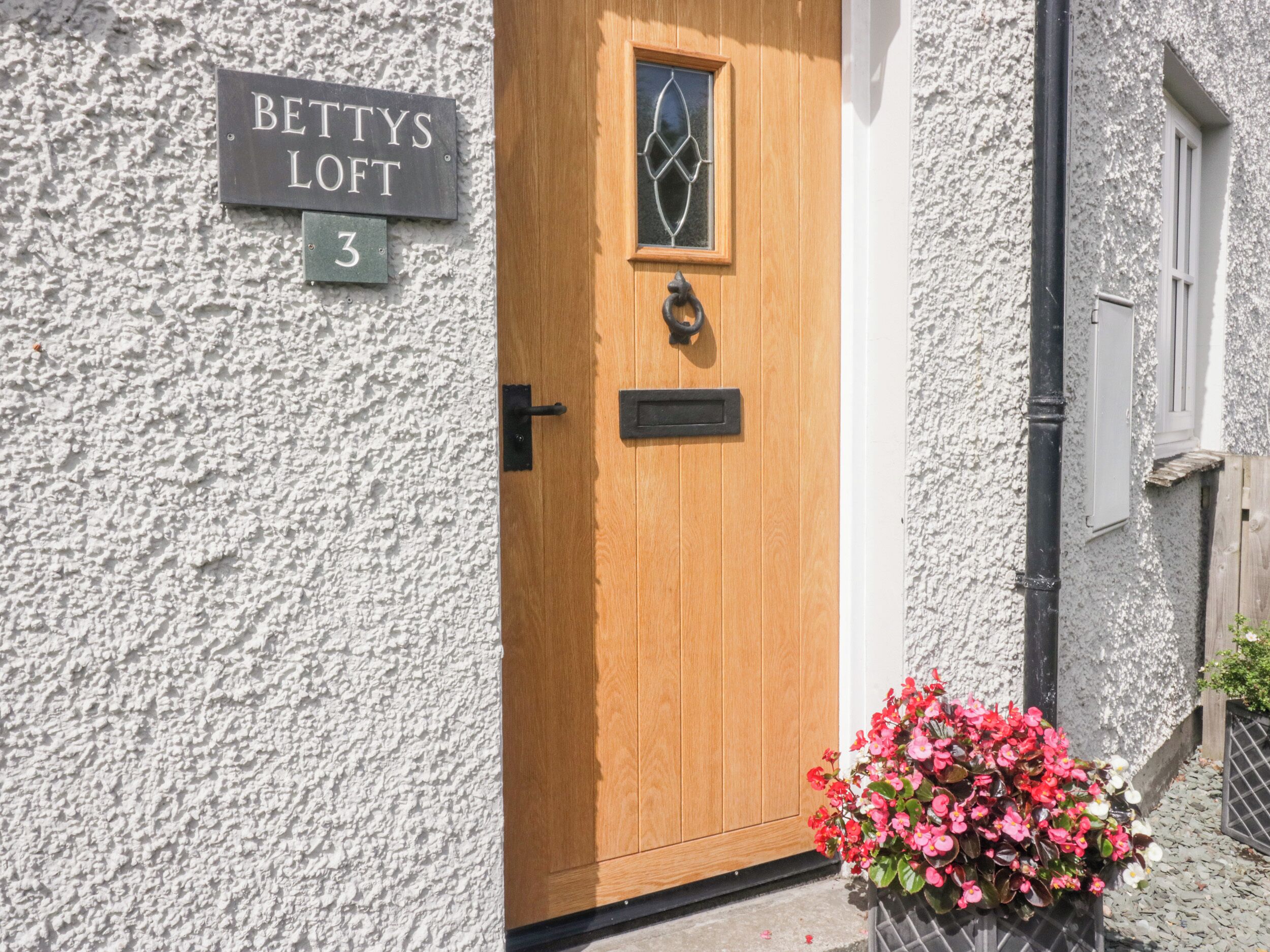 Una porta in legno con una finestra, un cartello sopra che recita "BETTYS LOFT 3" e un vaso con fiori rosa a terra.