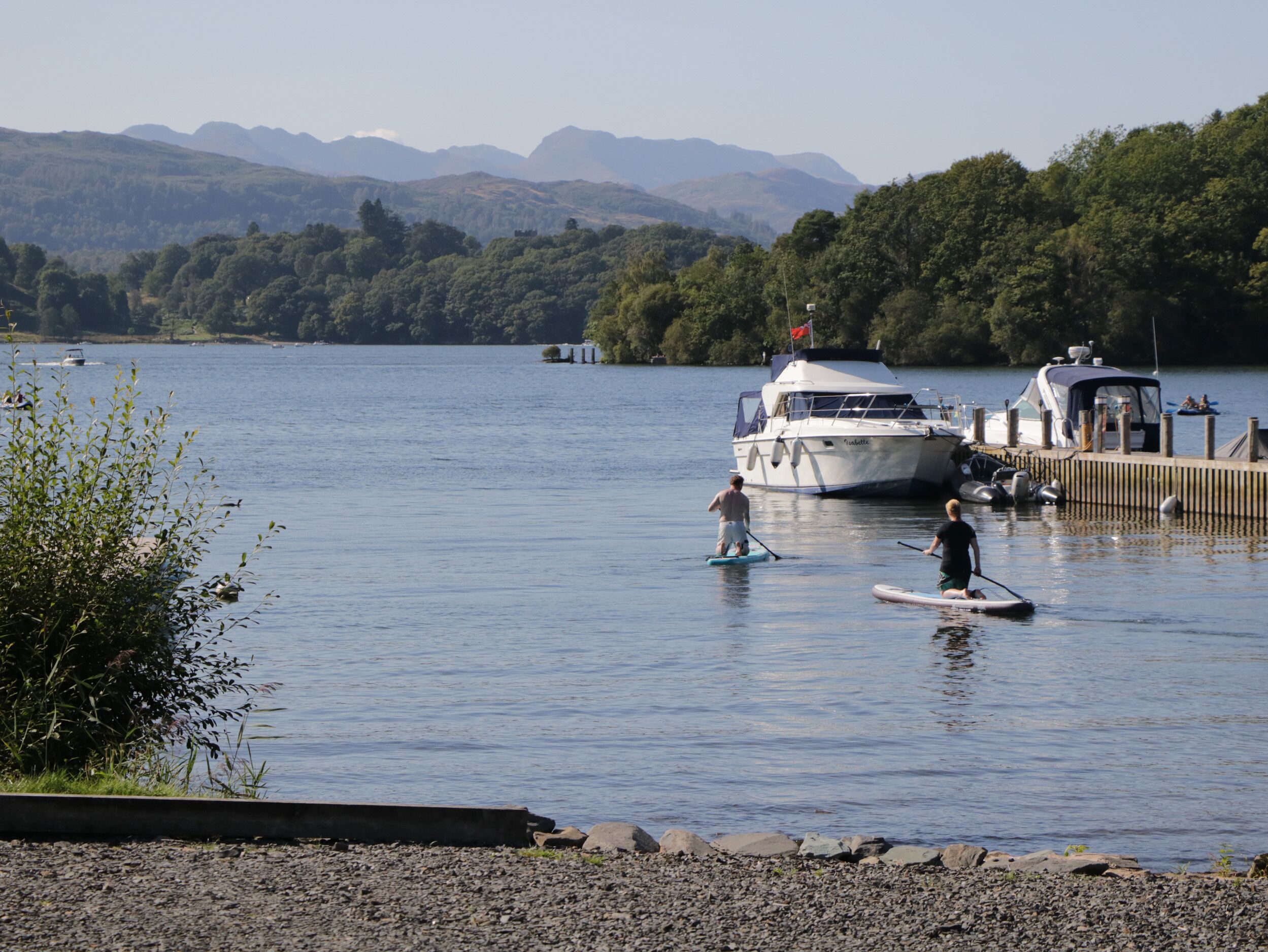 A calm lake with boats docked at a pier, surrounded by lush greenery and mountains in the background.