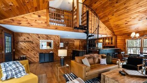 A cozy cabin interior with a wooden ceiling, a loft bedroom, a dining area, and a living room furnished with a sofa, armchair, and coffee table. - Quiet Cabin On Over 12 Acres Of Woods! (Becket)