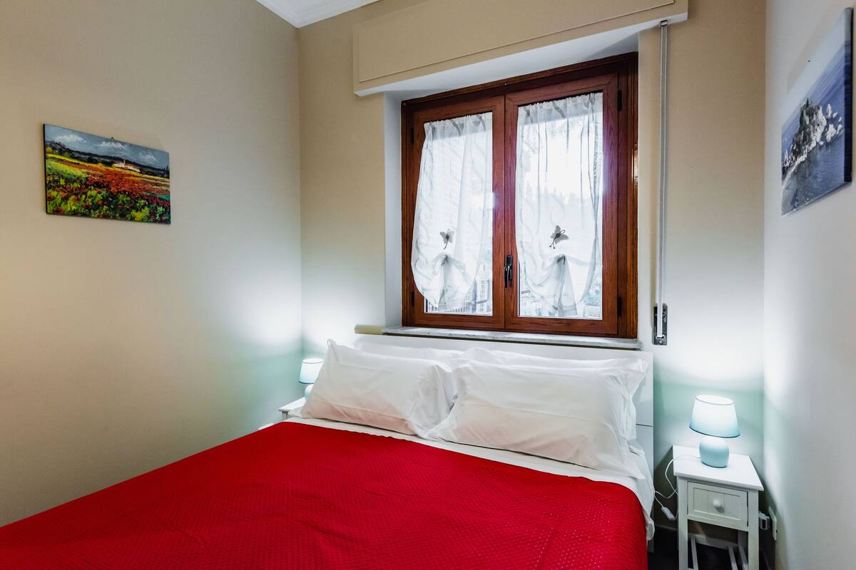 A bedroom with a red bedspread, white pillows, a window with curtains, a bedside lamp, and a painting on the wall.