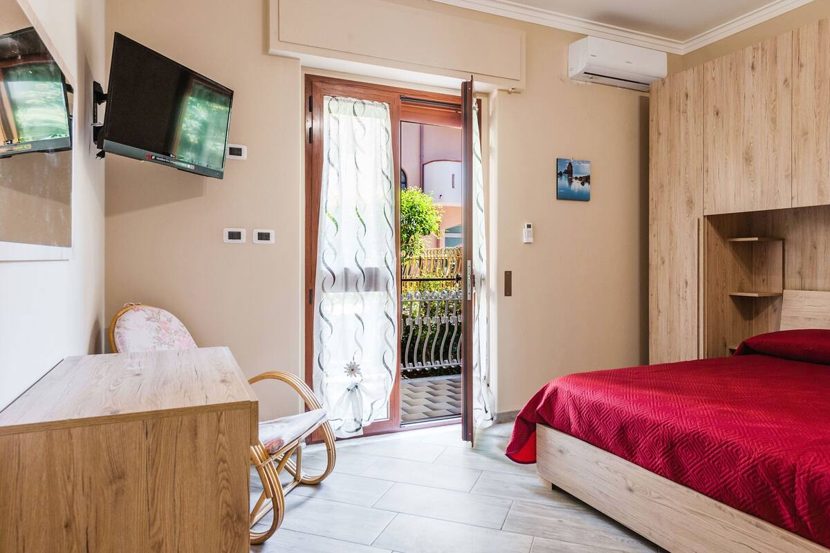 A bedroom with a bed, a desk, a chair, a TV mounted on the wall, and an open door leading to a balcony.