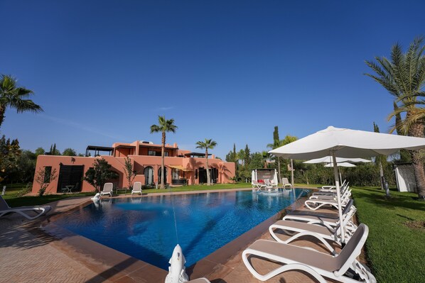 Una piscina con sillas de descanso y sombrillas, rodeada de palmeras y un edificio con fachada de estuco.