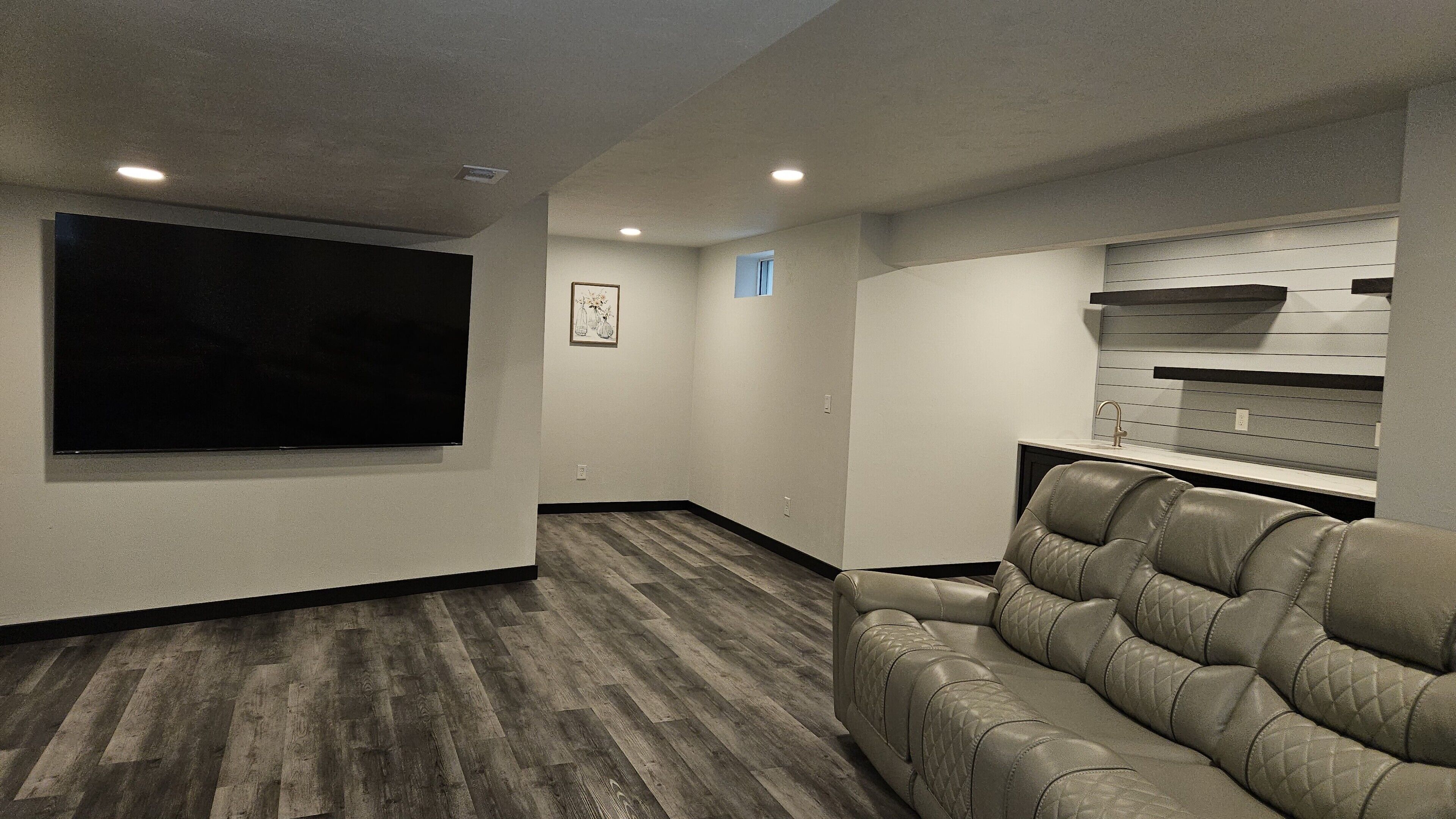 Un salón de cine en casa con un televisor de pantalla plana grande, un sillón de cuero reclinable y un mueble empotrado con un lavamanos.