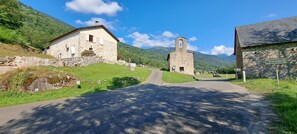 A rural road leading to a stone chapel with a bell tower, surrounded by stone buildings and lush greenery. - Emplacement Unique Offrant un Magnifique Panorama Face à la Chaîne Pyrénéenne (Ercé)