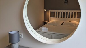 A bedroom with a round mirror, a bed with white bedding, a nightstand, and a lamp. - Hellaberg III: Quiet vacation home with sauna on the edge of the forest (Schleusegrund)
