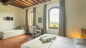 A bedroom with a bed, a desk, and a chair. There are windows with curtains, a television on the wall, and a view of the outdoors. - Podere La Quercia (San Gimignano)