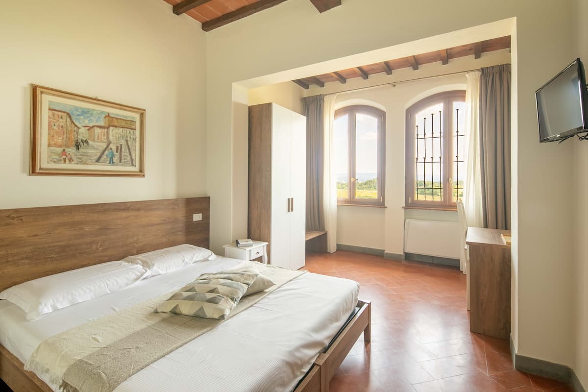 A bedroom with a wooden headboard, a large bed with pillows, a bedside table, a TV mounted on the wall, and a view of the outside through a window with curtains.