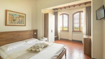 A bedroom with a wooden headboard, a large bed with pillows, a bedside table, a TV mounted on the wall, and a view of the outside through a window with curtains.