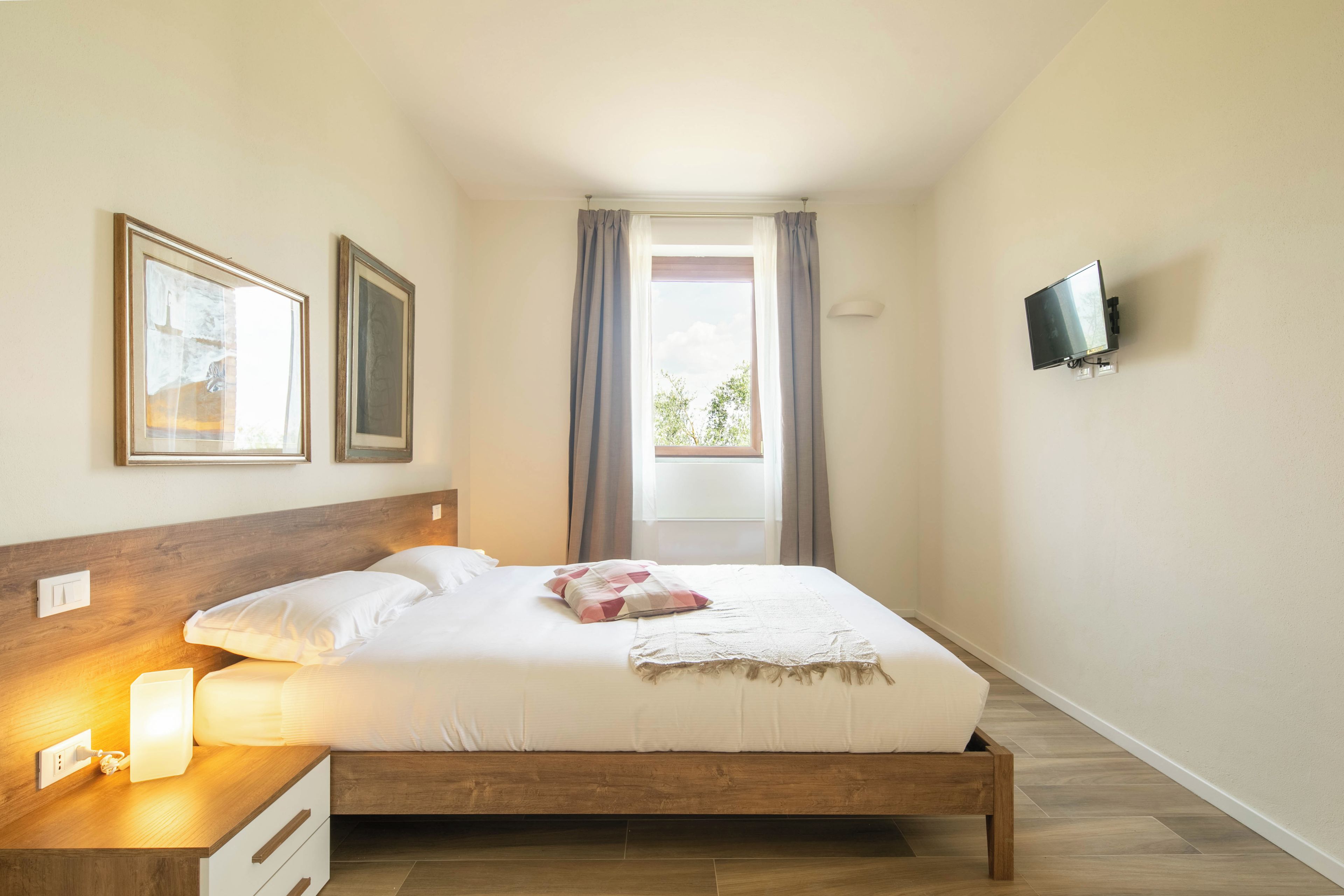 A neatly made bed with a wooden headboard, a bedside table with a lamp, and a television mounted on the wall.