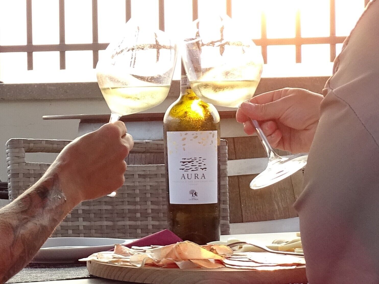 Una botella de vino con la etiqueta 'AURA' y dos copas sostenidas por una persona.