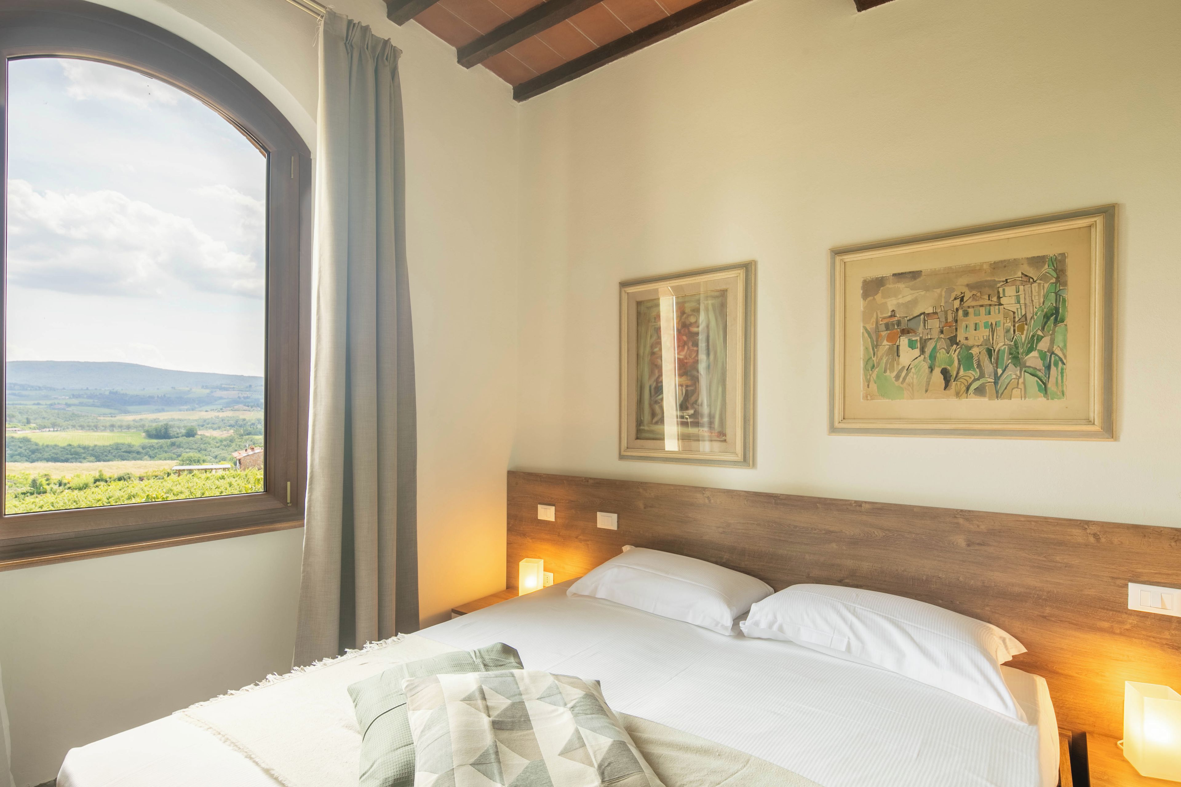 A bedroom with a large window, a bed with white linens, two wall-mounted lamps, and two framed paintings.