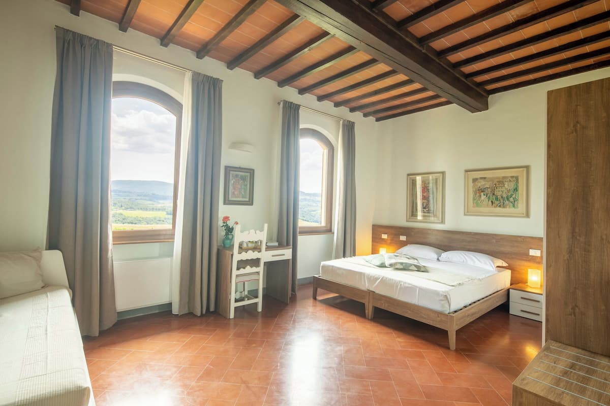 A bedroom with a bed, a desk, and a chair, large windows with curtains, and a view of the landscape.