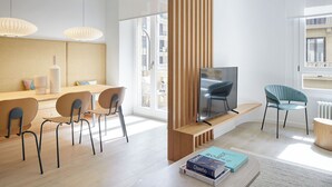 Uma sala de estar moderna com um suporte de TV de madeira, uma TV de tela plana, uma poltrona azul e um espaço de refeições com móveis de madeira e luminárias pendentes.