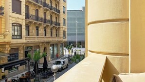 Uma varanda com tapete vermelho, com vista para uma rua com mesas ao ar livre e um edifício com varandas ornamentadas.