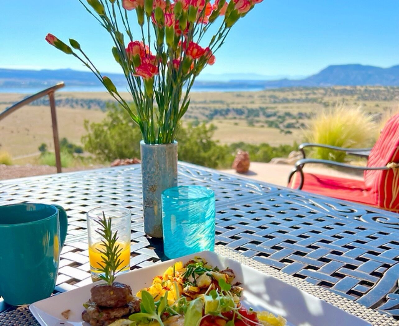 A dining table with a meal, a vase with flowers, and a view of a desert landscape.