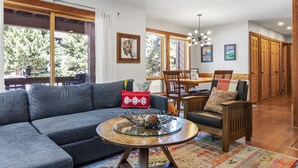 A modern living room with a sectional sofa, a glass coffee table, and a dining area with wooden furniture. - Outpost: Nez Perce C3 - Pool and Hot Tub Access! (Teton Village)