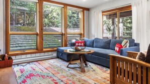 A cozy living room with a blue sofa, a wooden coffee table, and a colorful rug. Large windows provide a view of the outdoors. - Outpost: Nez Perce C3 - Pool and Hot Tub Access! (Teton Village)