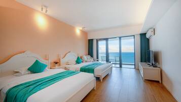 A hotel room with two beds, a desk, and a large window offering a view of the sea.