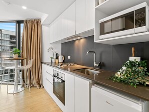 A modern kitchen with white cabinets, a stainless steel sink, and a microwave. There is a dining area with a table and chairs, and a plant on the counter.