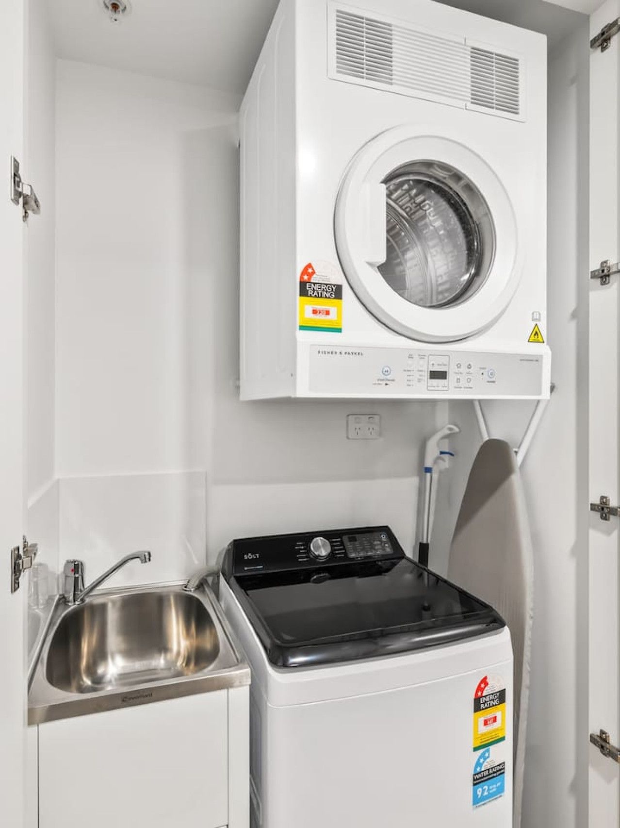 A compact laundry room with a front-loading washer and dryer, a stainless steel sink, and a safety warning label on the dryer.