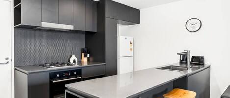 A modern kitchen with dark cabinetry, a stainless steel island, a built-in oven, and a refrigerator. There are two wooden stools at the island.