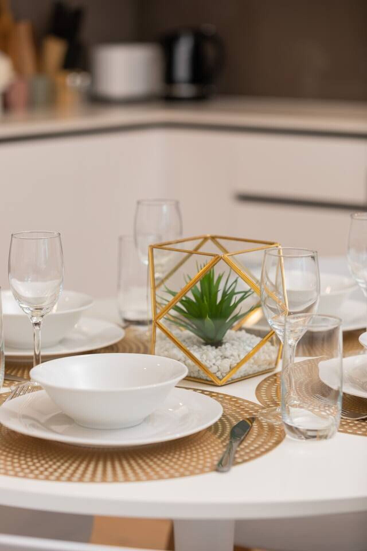 A dining table set with white plates, bowls, and clear glasses, centered around a decorative golden geometric frame with a small plant.