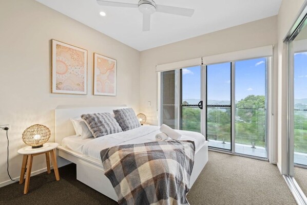 A bedroom with a bed, two bedside lamps, a ceiling fan, and a large window with a view.