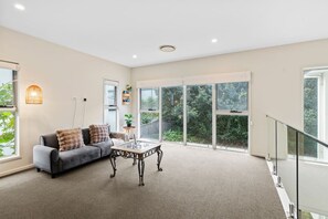 A spacious living room with a grey sofa, a glass coffee table, and large windows offering a view of greenery.