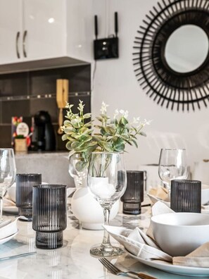 Una mesa de comedor dispuesta con vasos, un florero con follaje y un espejo moderno en la pared.