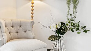 Ein geblueselter Sessel, eine Stehlampe, eine weiße Vase mit Pflanzen und ein Holzregal mit Grünpflanzen.